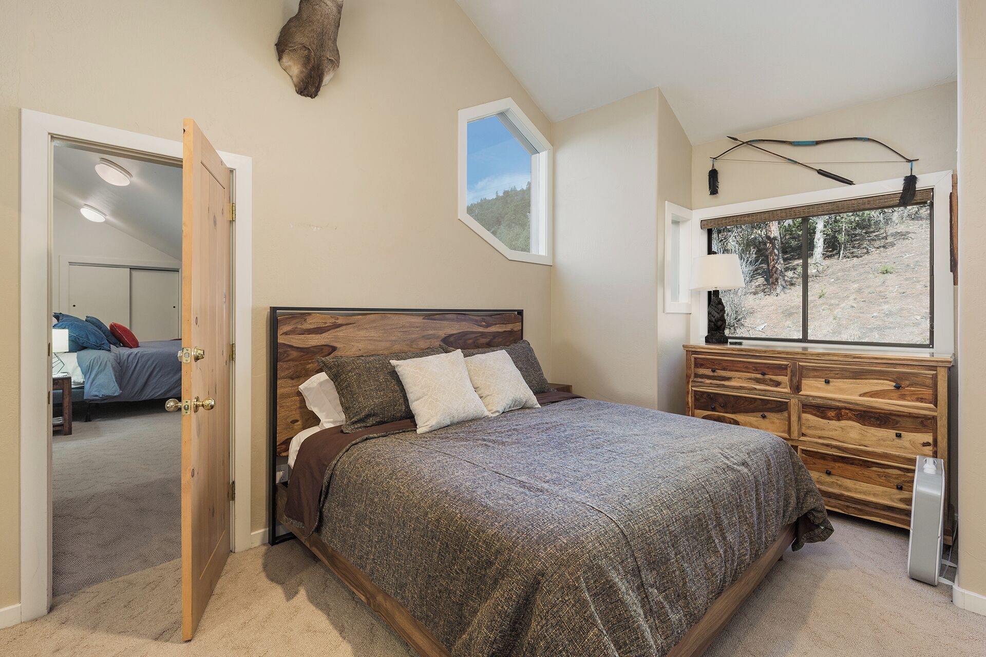 Camera da letto con un letto di legno, una cassettiera, una finestra vista alberi e una porta che conduce a un'altra stanza.