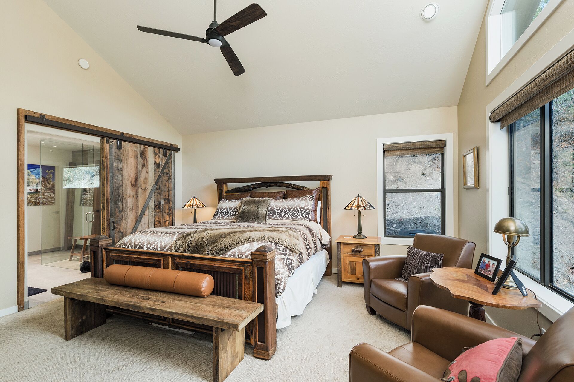 A spacious bedroom with a large bed, a wooden bench, a leather armchair, and a desk with a lamp.