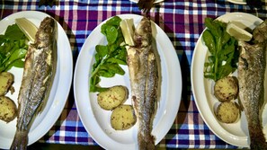 Three plates of grilled fish with potatoes and greens on a checkered tablecloth.