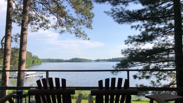 A deck with two wooden Adirondack chairs facing a lake, surrounded by trees. - Cozy lakefront chalet with cedar hot tub + stunning lake views (Remer)