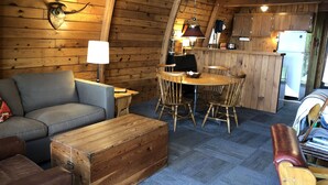 A cozy cabin interior with a wooden dining table, chairs, a sofa, and a leather ottoman. - Cozy lakefront chalet with cedar hot tub + stunning lake views (Remer)