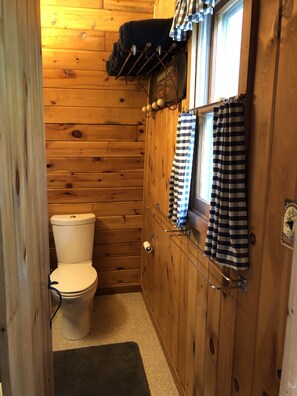 Een houten badkamer in een hutje, met een toilet, een raam met gordijnen en een plank met handdoeken.