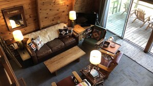 A cozy cabin interior with a wooden floor, a leather sofa, a wooden coffee table, a small wooden side table, a lamp, and a large window with a view of the deck. - Cozy lakefront chalet with cedar hot tub + stunning lake views (Remer)