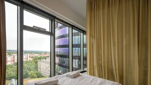 Een hotelkamer met een groot raam, een bed met witte lakens en uitzicht op gebouwen en groen.