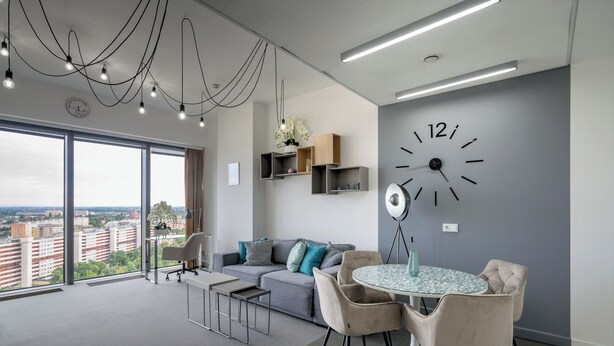 Een moderne woonkamer met een groot raam, een ronde tafel met stoelen, een bank en een wandklok.