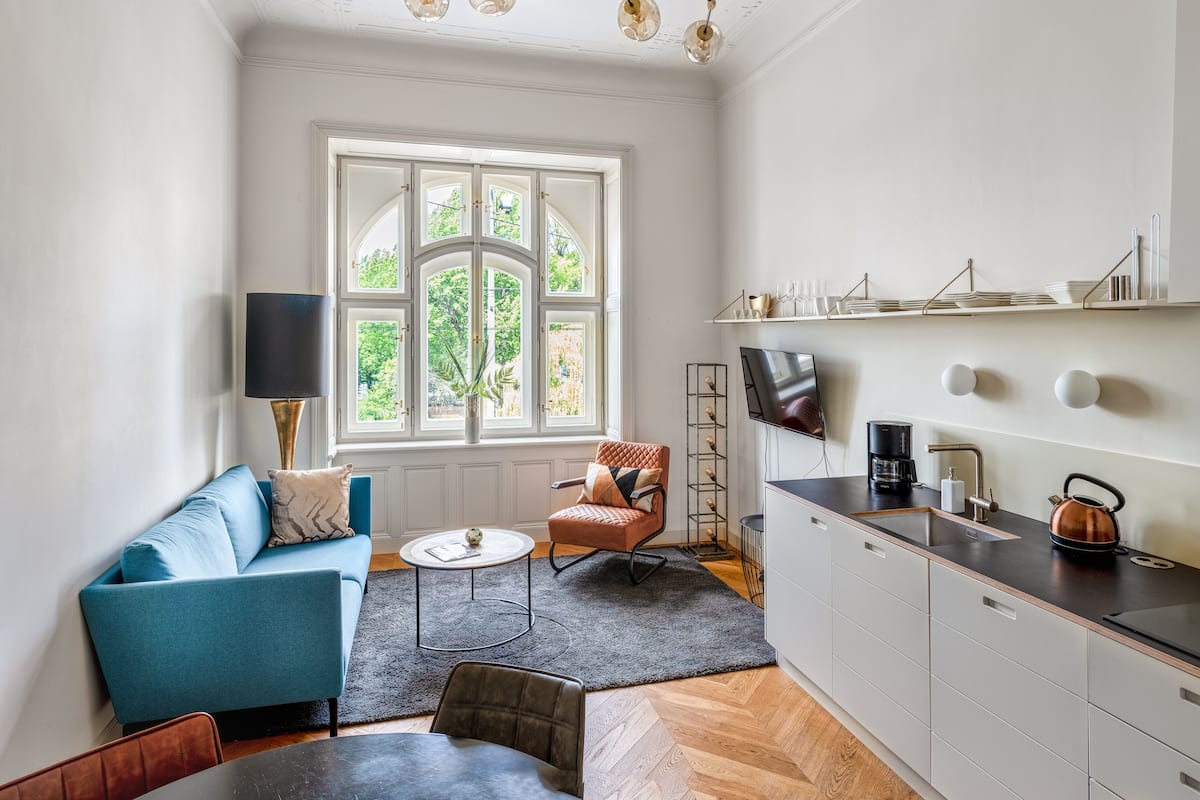 A modern living room with a blue sofa, a coffee table, a fireplace, and a kitchen area with a kettle and shelves.