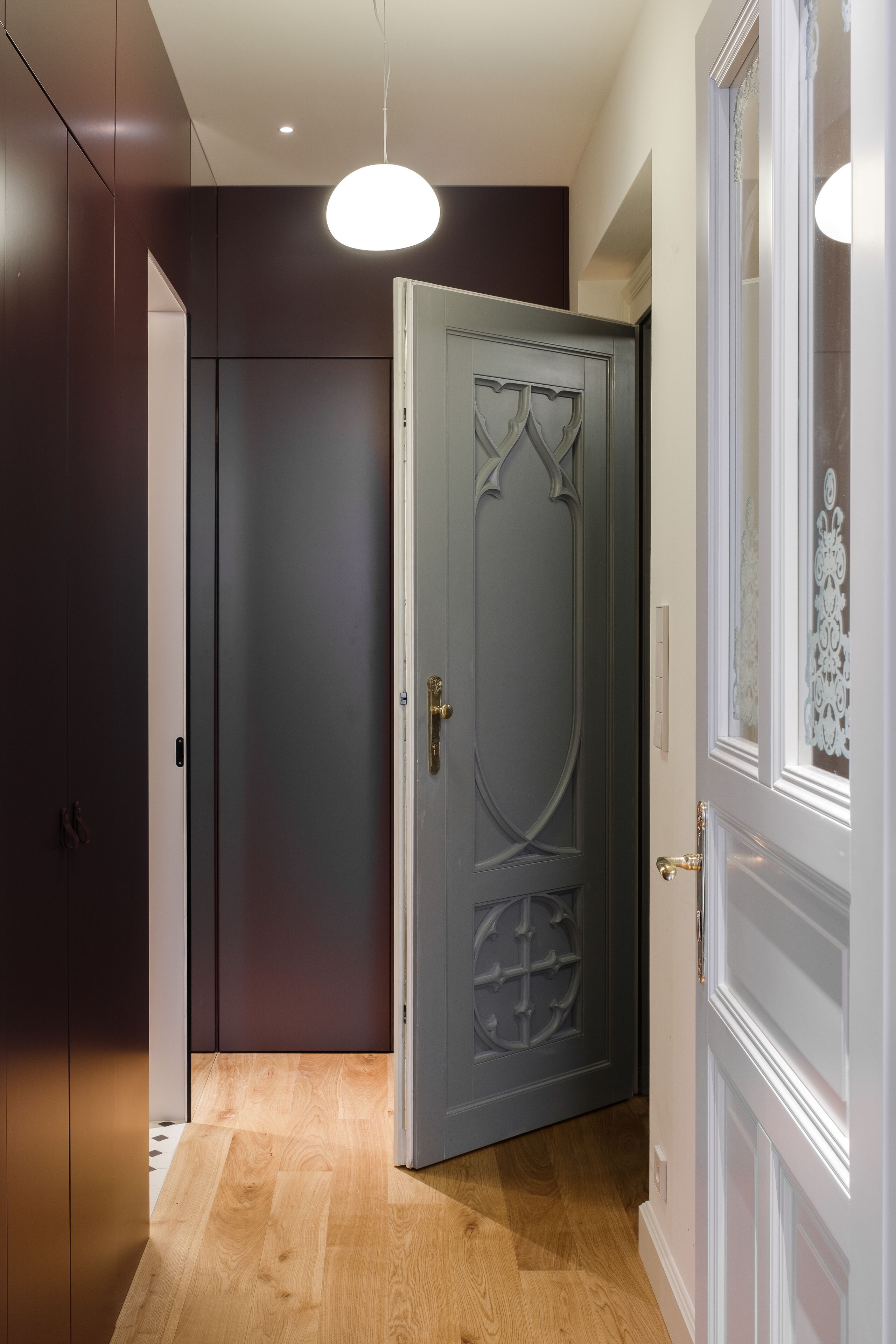 A hallway with a wooden floor, a dark door with a decorative panel, and a white door with a glass panel.