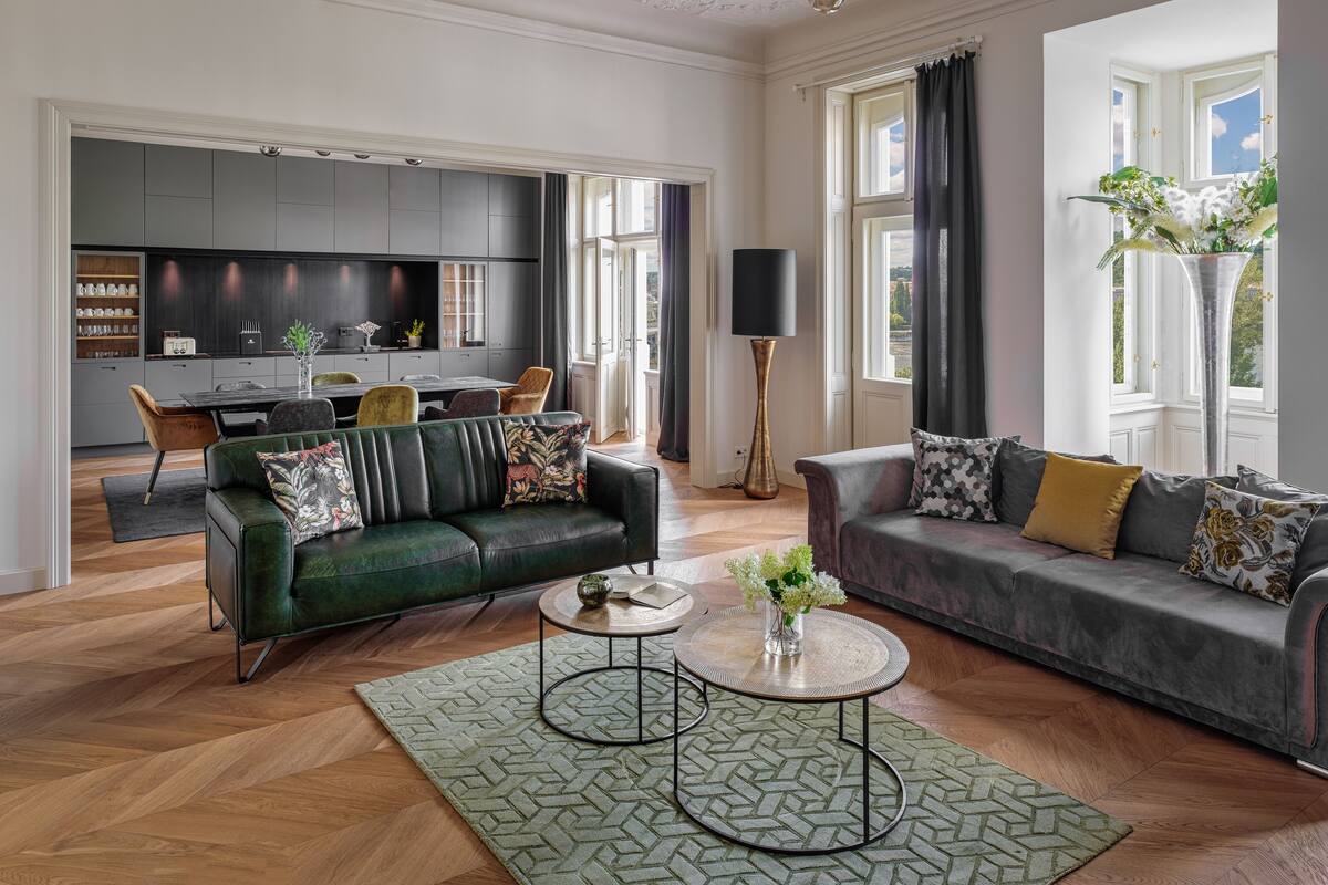 A spacious living room with a green sofa, a grey sofa, and a coffee table. There is a dining area with a table and chairs, and a kitchen in the background.