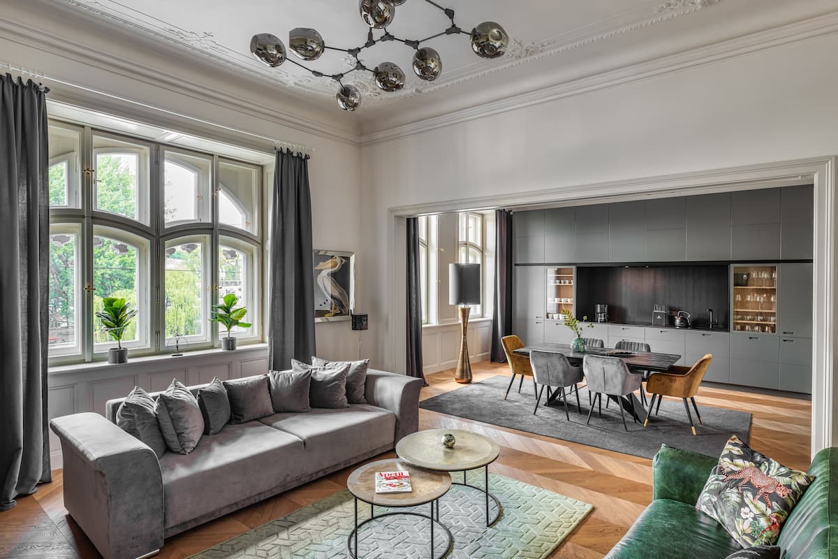 A spacious living room with a grey sofa, a dining area with a table and chairs, a kitchen with grey cabinets, and large windows with curtains.