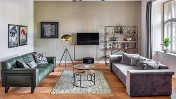 Ein modernes Wohnzimmer mit einem grünen Sofa, einem grauen Sofa, einer Stehlampe, einem Couchtisch und einem an der Wand montierten Fernseher.
