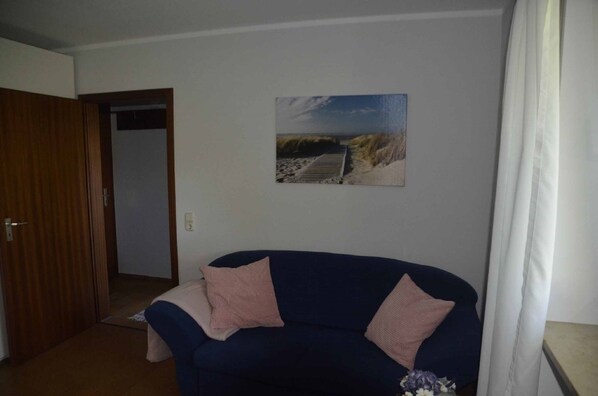 A room with a dark blue sofa, two pink cushions, a framed picture of a beach scene, and a wooden door. - Ferienwohnung Biedermann (Schoenwald)