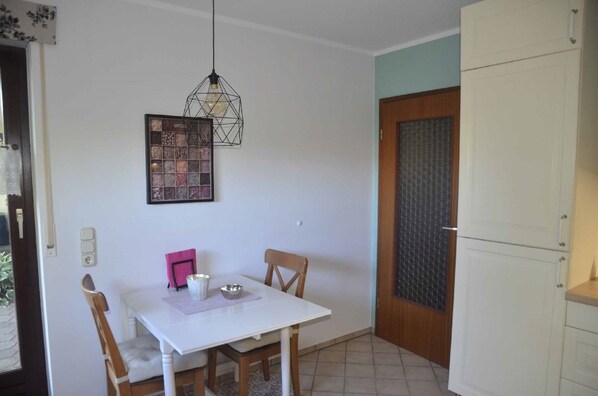 A small dining area with a white table, two chairs, a hanging light fixture, and a door with a patterned glass panel. - Ferienwohnung Biedermann (Schoenwald)