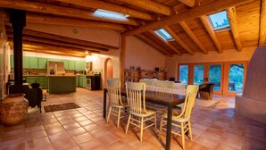 A rustic kitchen with wooden beams, a dining area with a table and chairs, and a view of greenery through large windows. - Peaceful family/group home with private attached treehouse (Canones)