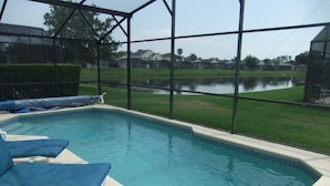 A swimming pool with blue cushioned chairs, surrounded by a covered area with a view of a lake and houses in the background. - Ref 03 Luxury 4 Bed Villa With Lake View own Pool (Kissimmee)