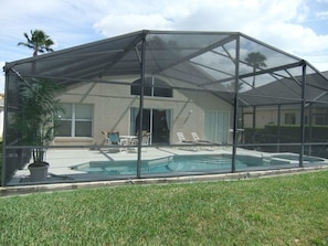 A covered outdoor area with a pool, a house in the background, and a lawn.