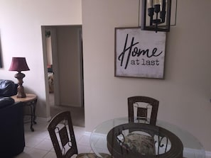 A dining area with a glass table, wicker chairs, and a sign that reads "Home at last" on the wall.
