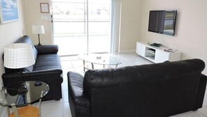 Ein modernes Wohnzimmer mit einem schwarzen Ledersofa, einem Glastisch, einer weißen TV-Konsole und einem großen Fenster mit Blick ins Freie.