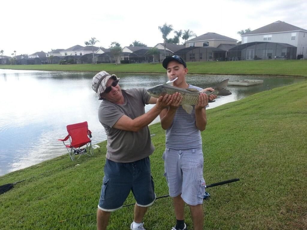 Two people holding a fish near a body of water with houses in the background.