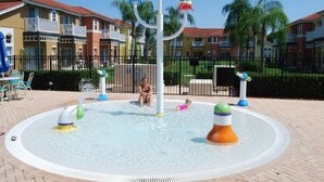 Ein Wasserspielplatz mit einer zentralen Säule und bunten Wasserstrahlen, umgeben von einem gepflasterten Weg und Wohnhäusern im Hintergrund.