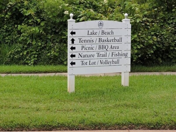 Un cartel direccional con flechas indicando diversas actividades recreativas como lago/playa, tenis/baloncesto, área de picnic/barbacoa, sendero natural/pesca y parque infantil/voleibol.