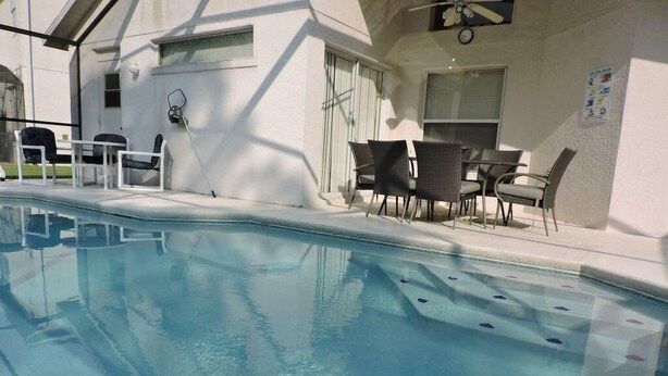 A swimming pool with a sunshade and chairs, surrounded by a building with a white exterior and a clock on the wall.