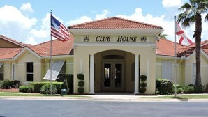 A building with a sign reading "CLUB HOUSE" at the entrance, featuring a red-tiled roof, a flag, and a palm tree.