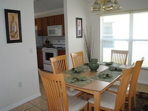 A dining area with a wooden table, four chairs, and a green placemat. There is a large window with blinds, a kitchen in the background, and a framed picture on the wall.