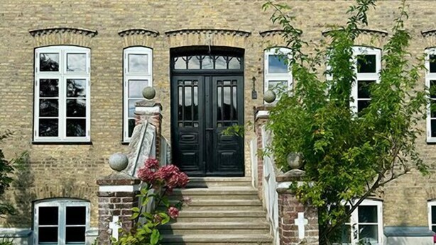 Un bâtiment en pierre de deux étages, avec une porte d’entrée noire, de nombreuses fenêtres et un petit jardin agrémenté d’une plante en pot.