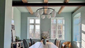 Une salle à manger avec une table en bois, des chaises et un lustre. Il y a des fenêtres avec des cadres blancs, et de là, on peut voir les arbres à l’extérieur.
