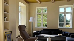 Un salon avec un fauteuil en cuir marron, un canapé noir, un pouf jaune et une lampe blanche. On y trouve des étagères et des fenêtres donnant sur un paysage verdoyant.