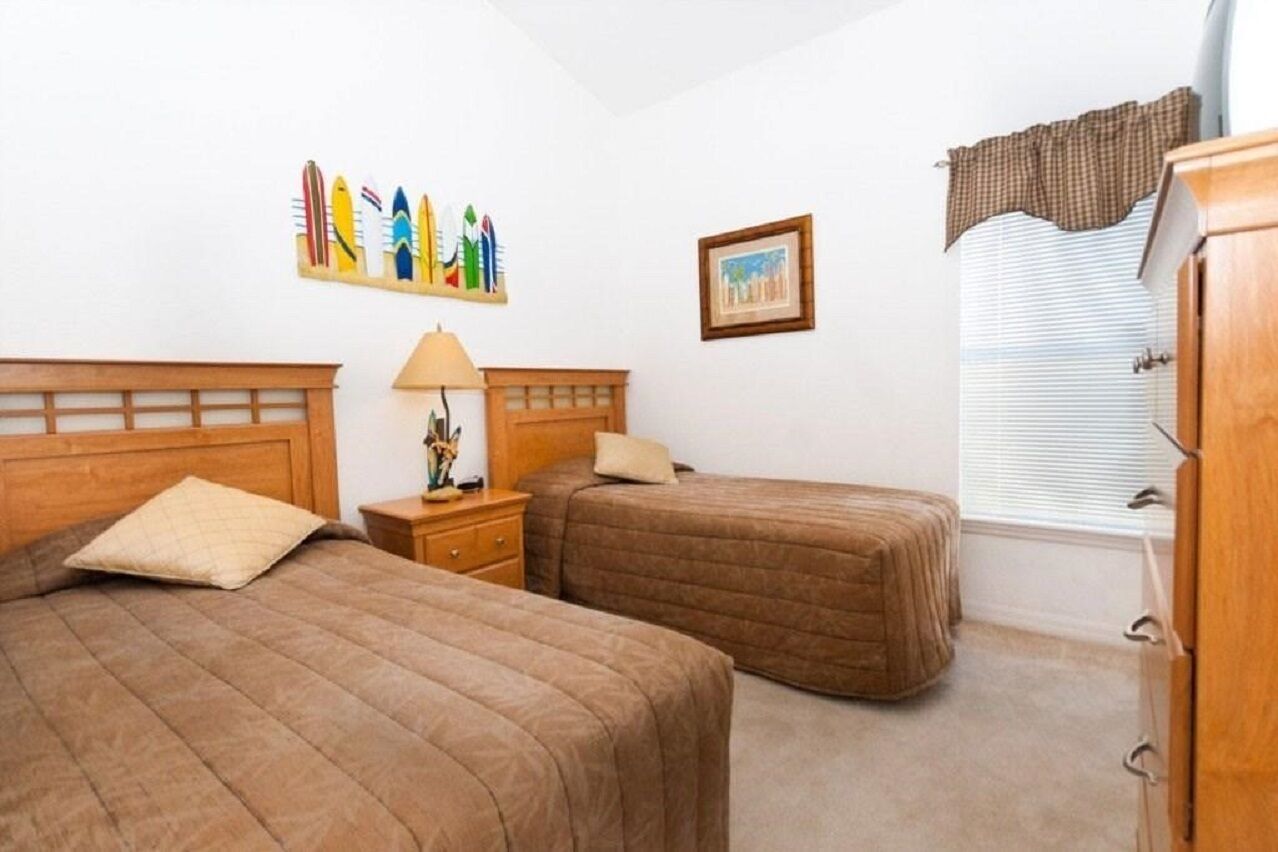 A room with two beds, a nightstand, a lamp, a window with blinds, and a wall with artwork.