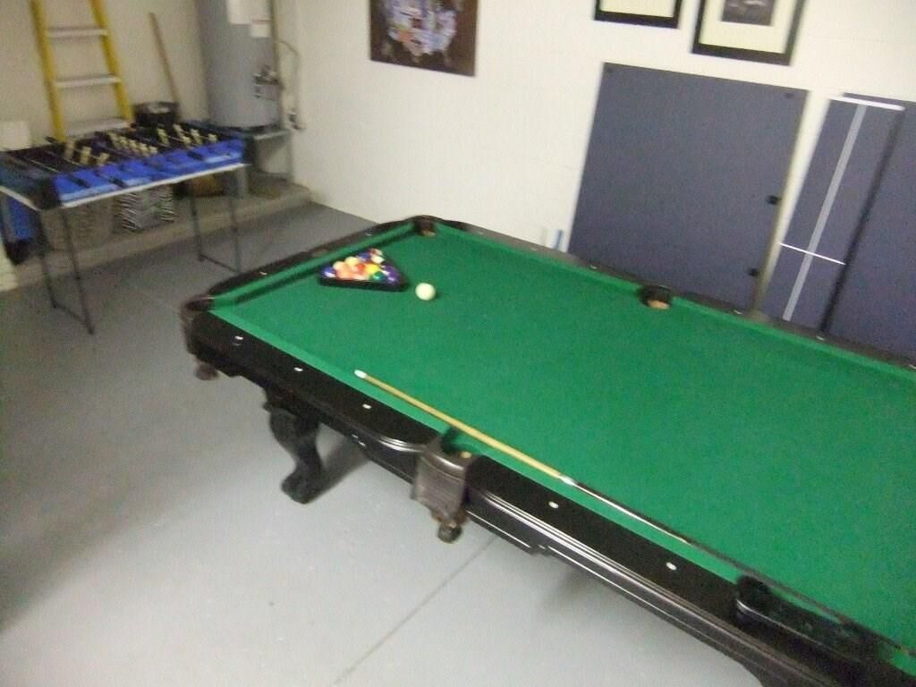 A pool table with a green felt surface, a cue ball, and a set of pool cues. There are blue storage bins to the left and a ladder against the wall.