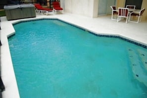 A swimming pool with a tiled edge, surrounded by white lounge chairs and a covered hot tub.