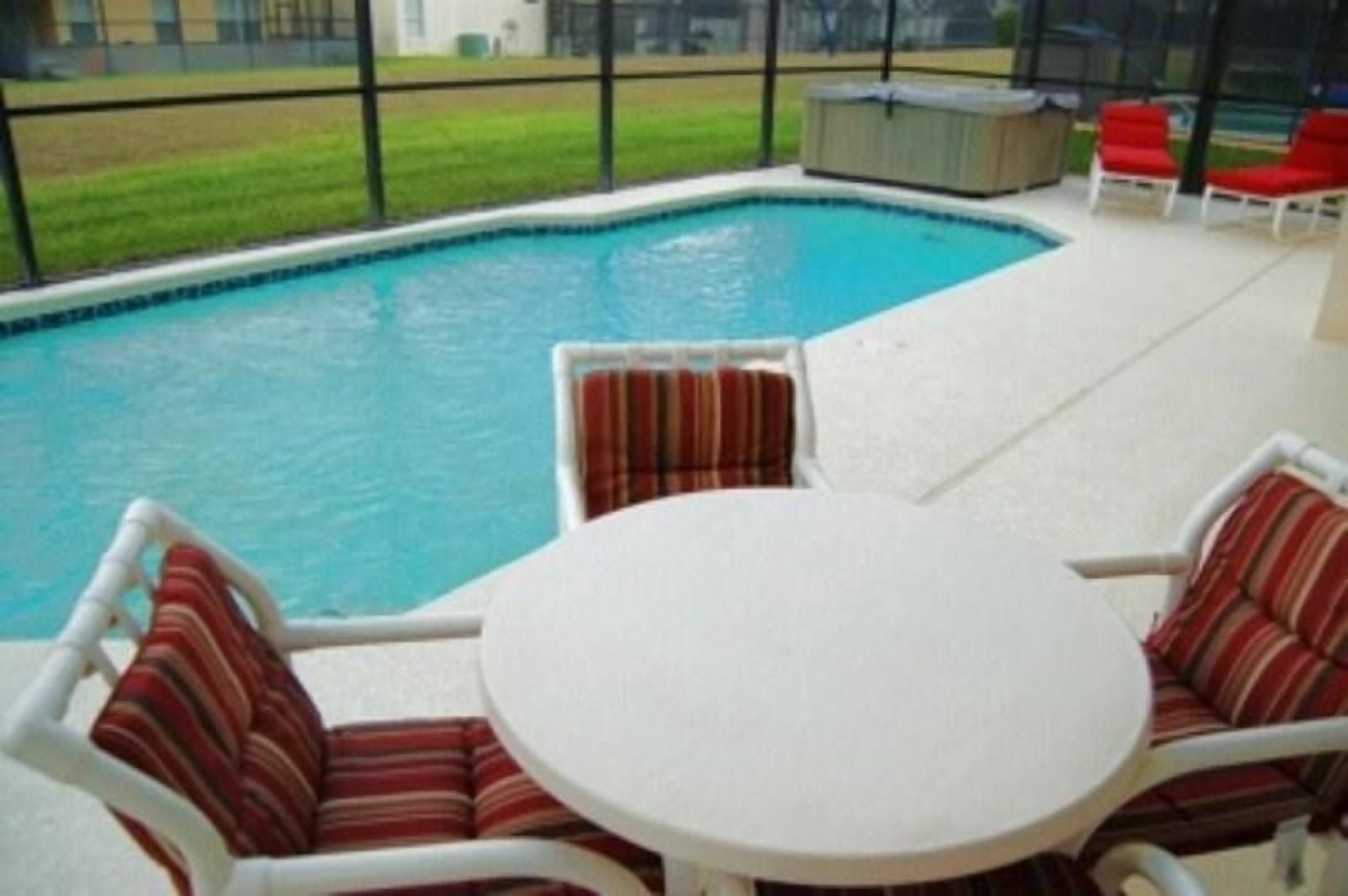 A pool area with a round table and two chairs, a hot tub, and a covered seating area with red chairs.