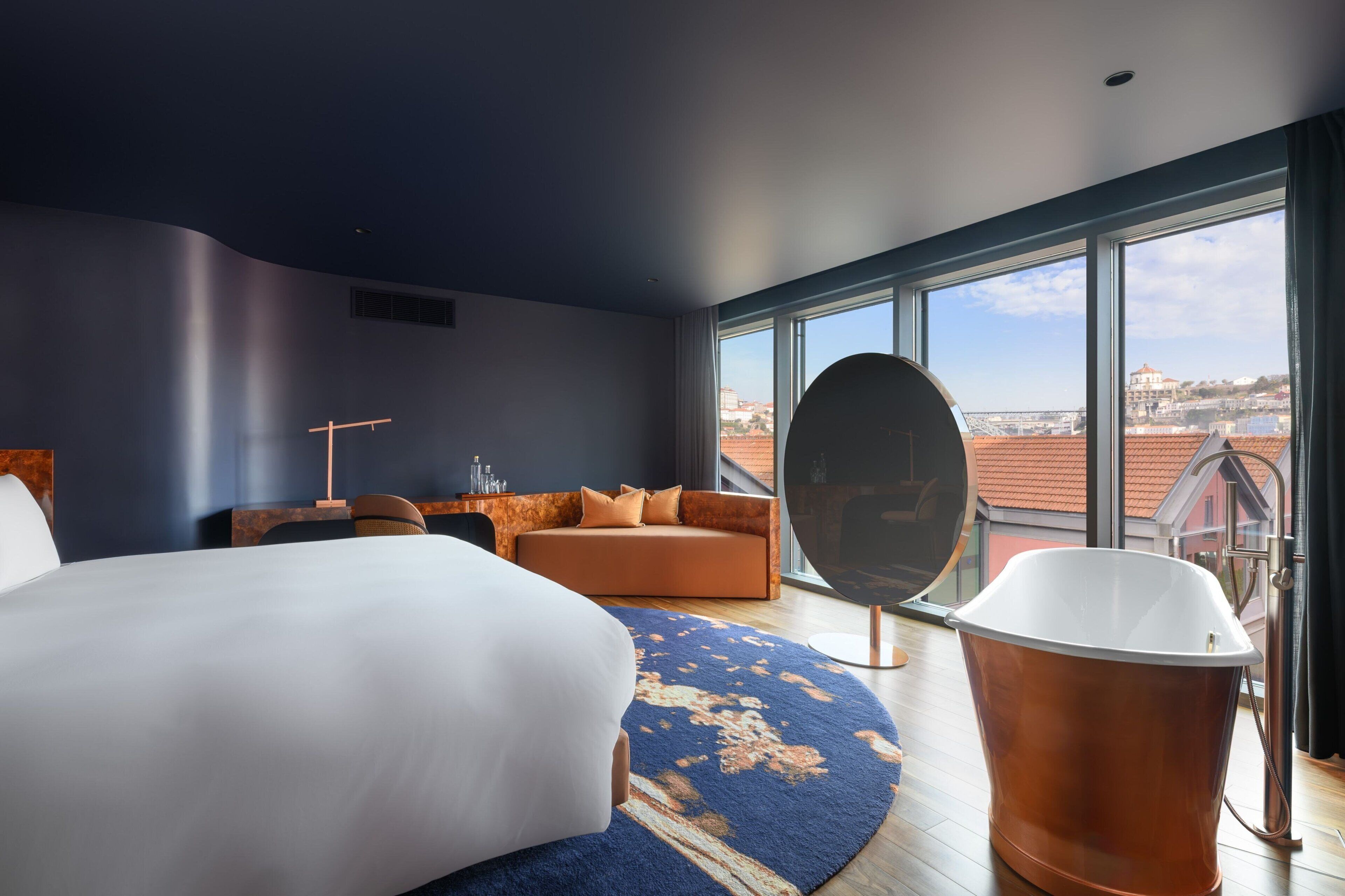Un hotel moderno con una cama grande, un espejo redondo, una bañera y vistas a la ciudad a través de ventanas amplias.