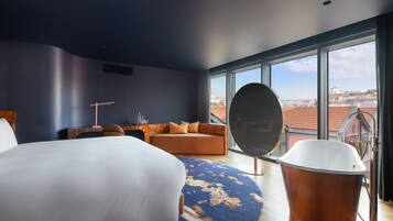Un hotel moderno con una cama grande, un espejo redondo, una bañera y vistas a la ciudad a través de ventanas amplias.