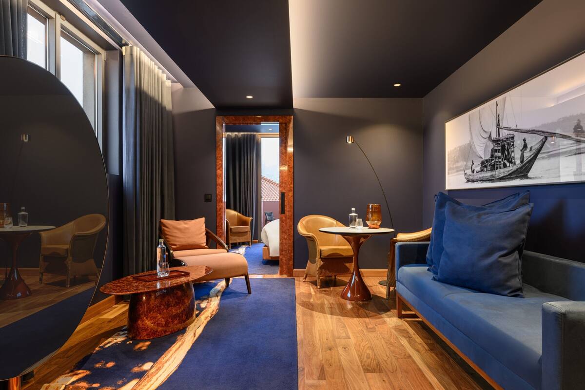 Une chambre d’hôtel moderne avec un canapé bleu, une table basse en bois, une petite table ronde et une grande fenêtre avec des rideaux.