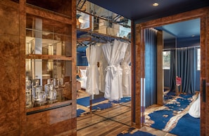 A hotel room with a large mirror, a wooden cabinet with glassware, a blue carpet, and white robes hanging on a rack.