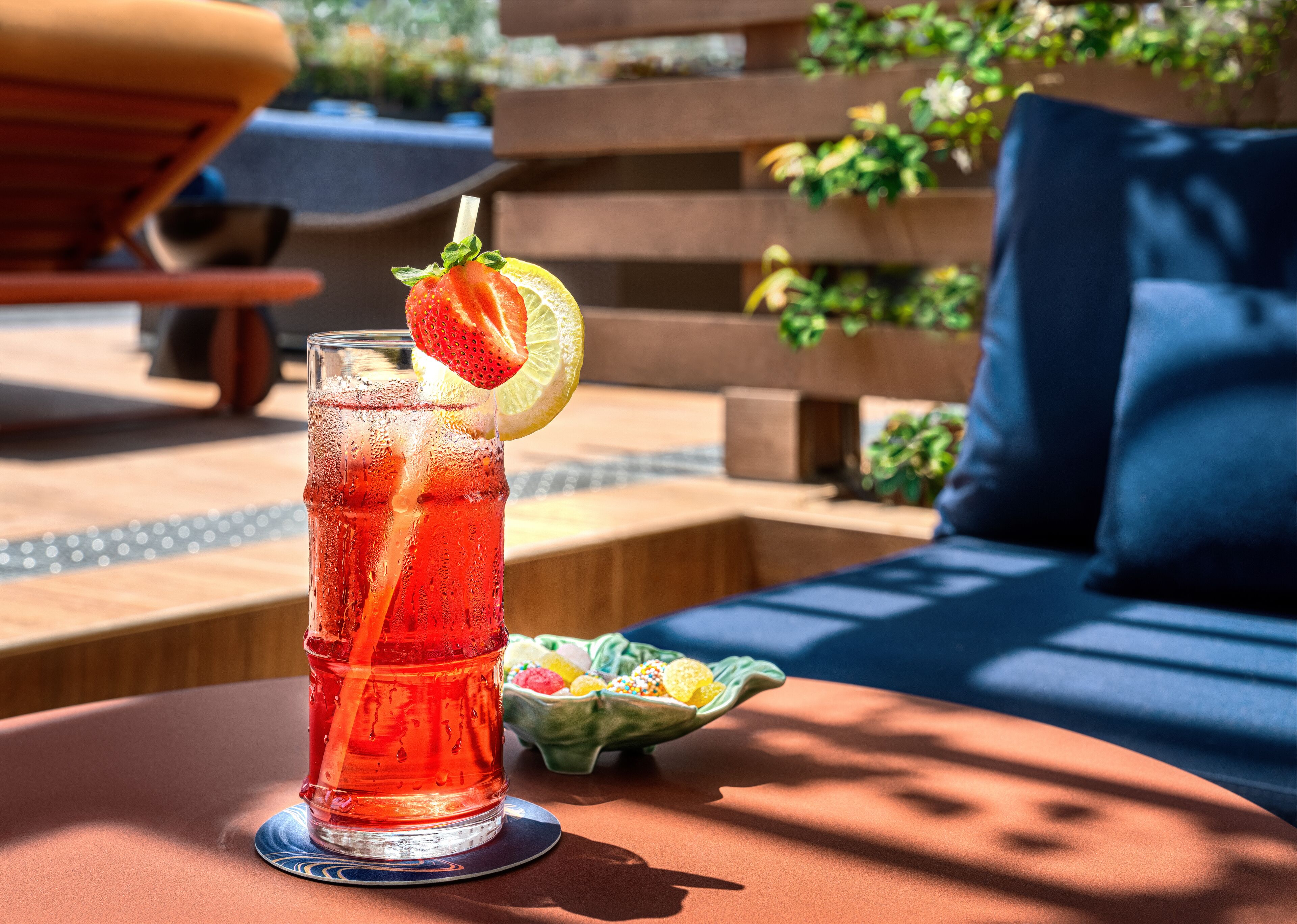Un verre d’une boisson rouge, garni d’une tranche de fraise et d’une tranche de citron, est posé sur une table à l’extérieur.