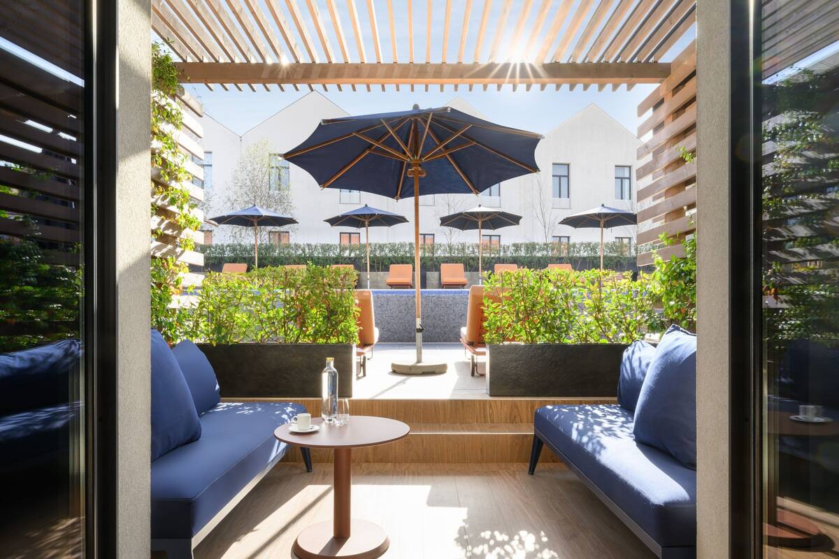Un espace extérieur aménagé avec des canapés bleus et des tables rondes, entouré de plantes en pot et de parasols.
