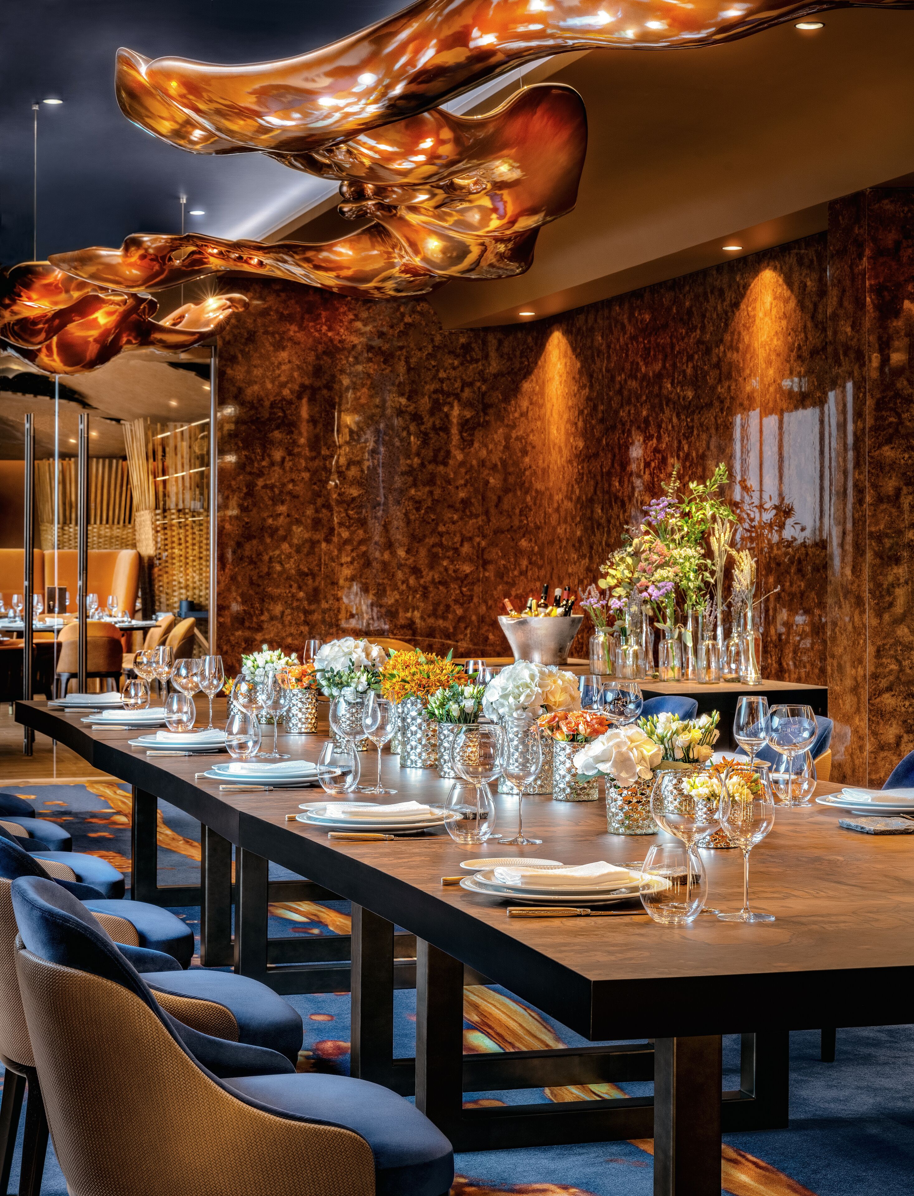 Una larga mesa de comedor dispuesta con copas y centros de mesa florales, rodeada de sillas tapizadas.