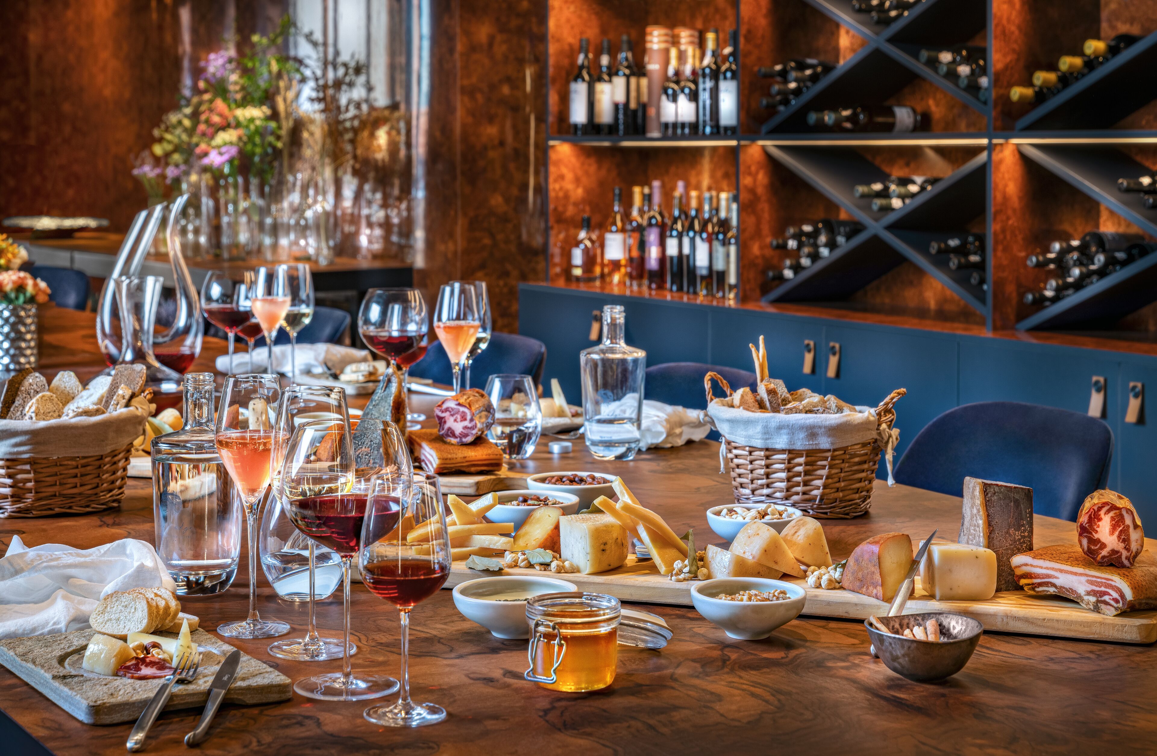 A well-set dining table with various cheeses, breads, and wine glasses, surrounded by a wine rack and baskets.