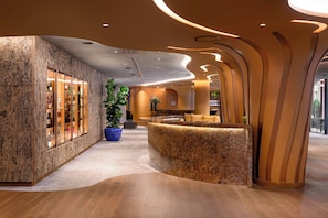 A modern lobby with a reception desk, a large potted plant, and a stone wall with glass display cases.