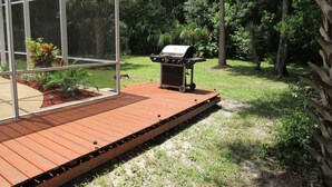 A wooden deck with a barbecue grill, surrounded by lush greenery and a glass enclosure. - Ref 24 Modern 4 Bed Villa With own South Facing Pool (Kissimmee)