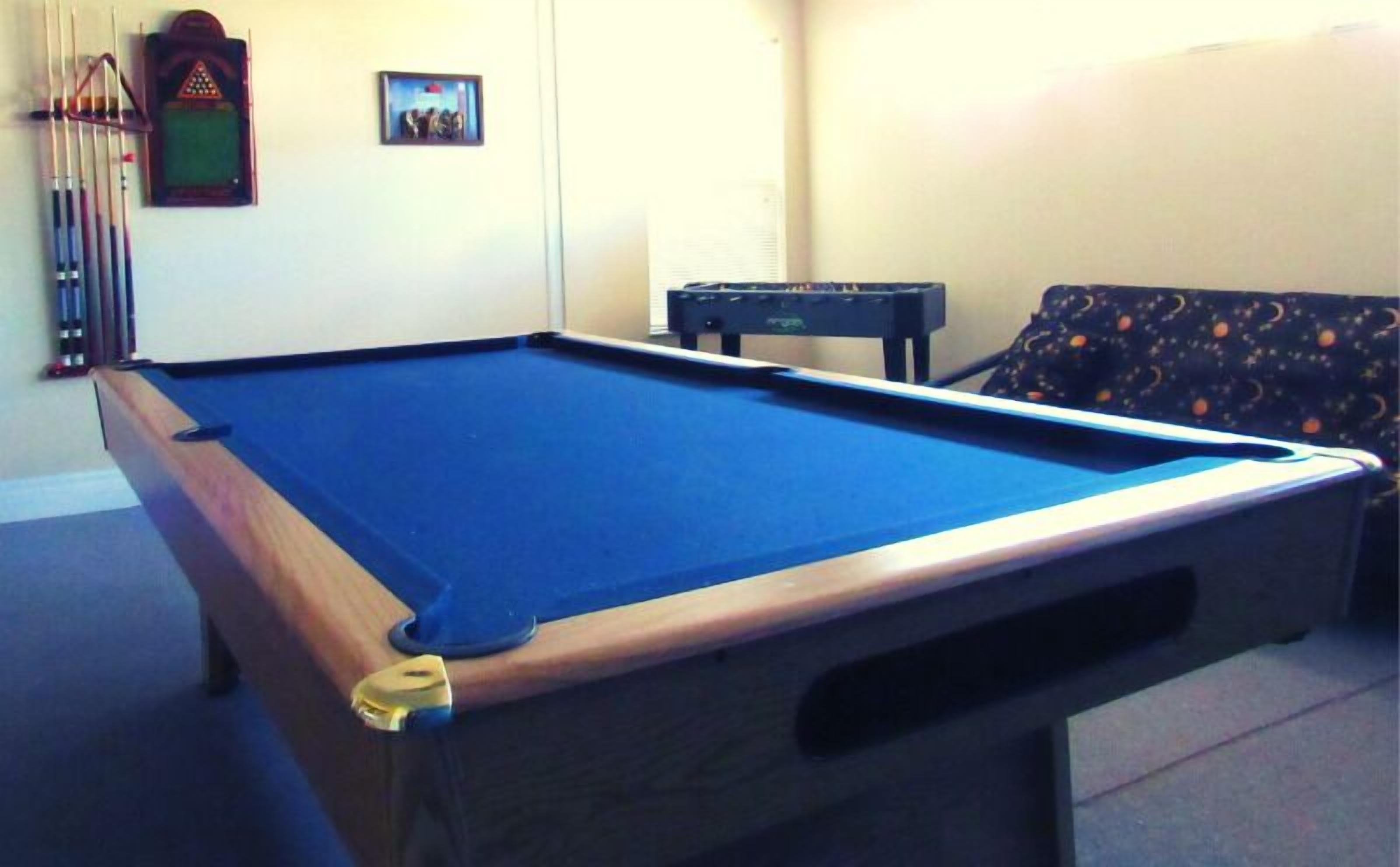 A pool table with a blue felt surface, a wooden side rail, and a pocket. There is a pool cue rack against the wall and a small table with a dark cloth.