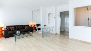 Een moderne woonkamer met een glazen salontafel, een zwarte bank met bruine kussens, en een eethoek met een glazen tafel en stoelen.