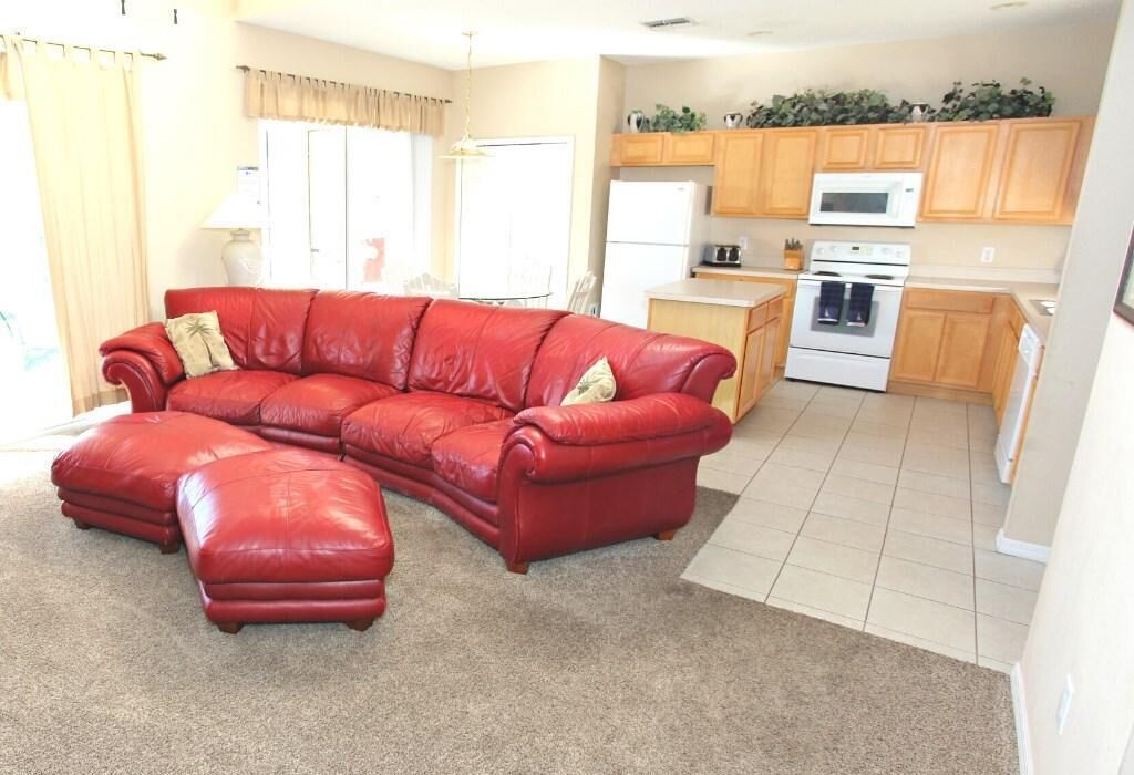 A living room with a red leather sectional sofa and ottoman, a kitchen with wooden cabinets and appliances, and a large window with curtains.
