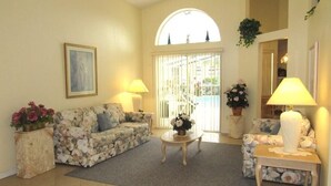 A living room with a floral sofa, a round coffee table, and two lamps. There is a painting on the wall and a window with a view of the outside.