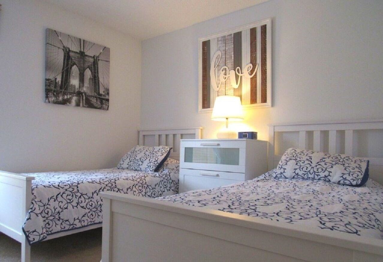 Ein Schlafzimmer mit zwei Einzelbetten, einem Nachttisch mit Lampe, einem an der Wand hängenden Bild einer Brücke und einem Schild mit der Aufschrift 'Love' über dem Bett.