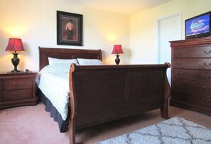 A bedroom with a wooden bed, two bedside tables with lamps, a dresser, a framed picture on the wall, and a television on the dresser.
